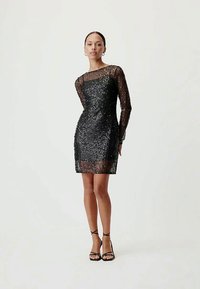 Mulher vestindo um vestido mini preto de manga longa e lantejoulas com sobreposição de tecido transparente, combinado com sandálias de salto alto pretas e com tiras, em pé.