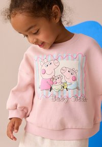 Niña sonriente con un sudadera rosa con el dibujo de la familia Peppa Pig y el texto "Hermana Bebé", frente a un fondo neutro con una silla azul.