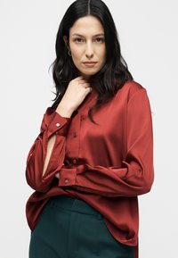 Blouse en satin rouge avec manches longues, poignets boutonnés et boutons ton or. Portée avec un pantalon vert. Texture lisse, coupe décontractée.