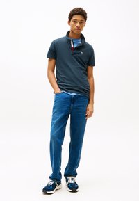 Ung mann som står med den ene hånden i lomma, iført marineblå polotrøye, lyseblå undertrøye, blå jeans og blå joggesko på hvit bakgrunn.