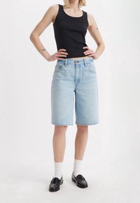 Henkilö, joka on pukeutunut vaaleansinisiin polvipitkiin farkkushortseihin, mustaan t-paitaan, valkoisiin sukkiin ja mustiin loafereihin, seisoo kädet lanteilla.