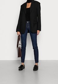 Blazer negro oversized, top negro de canalé, jeans ajustados azules oscuros y mocasines negros. Bolso de mano en relieve de cocodrilo marrón como accesorio.