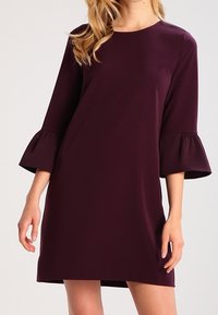 Un vestido de longitud a la rodilla, de color ciruela, con un escote redondo y mangas tres cuartos en forma de campana, hecho de una tela suave.