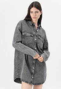 Grå denim skjortekjole med knapfront, to brystlommer og lange ærmer. Har en afslappet pasform og en rundet søm.
