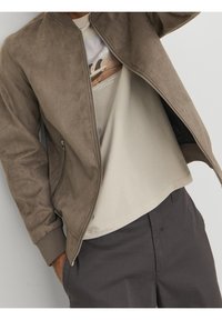 Taupe bomberjack in een zacht, suede-achtige stof met ribgebreide boorden, een zilveren rits en zijzakken. Gecombineerd met een beige t-shirt en donkere broek.