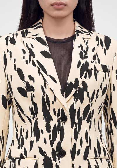 Femme portant un blazer crème avec des taches d'imprimé animalier noir irrégulier et des boutons noirs sur un haut sombre transparent.