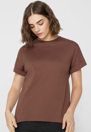 Žena s vlnitými hnědými vlasy po ramena, oblečená v jednobarevném hnědém tričku s krátkým rukávem, dívající se dolů s uvolněným postojem.