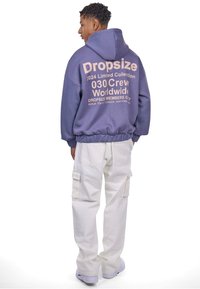 Lilla kapuutsi, mille seljal on heleda roosa teksti print "Dropsize 2024 Limited Collection", koos valgete cargo-pükste ja sobivate kingadega.