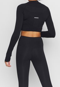 Schwarzes, figurbetontes Sportcrop-Top mit langen Ärmeln, strukturiertem Einsatz und reflektierenden Details auf dem Rücken, kombiniert mit passenden hochgeschnittenen Leggings.