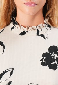 Primo piano della parte bassa del viso e del collo di una persona che indossa un top beige con motivi floreali neri e un colletto arricciato.