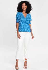 Blaues Wickeloberteil mit kurz rüschierten Ärmeln, Peplum-Taille und glatter Textur, kombiniert mit weißen Hosen mit fransigem Saum und Riemchen-High Heels.