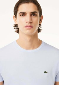 Camiseta de manga corta azul claro hecha de algodón, con un pequeño logo de cocodrilo bordado en verde en el lado izquierdo del pecho. Textura suave.
