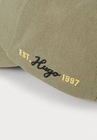 Olivengrøn kasket med kordfløjsstruktur, prydet med broderet "EST. Hugo 1997" i sort og gult. Krummede skygge og struktureret design.