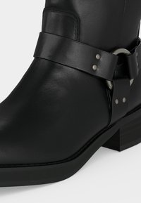 Μαύρο δερμάτινο μποτάκι αστραγάλου με χαμηλό τετράγωνο τακούνι και διακοσμητικό λουράκι με μεταλλικό κρίκο και καρφιά γύρω από τον αστράγαλο.