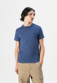 Tricou din bumbac albastru, cu mâneci scurte, decolteu rotund și un mic logo negru pe piept. Țesătura pare netedă și ușoară.