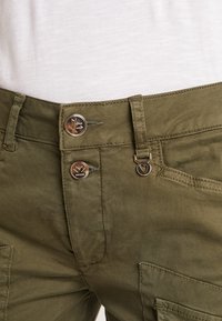 Gros plan sur un pantalon vert olive avec deux boutons métalliques et un pendentif en forme de cœur près de la poche droite, porté avec un haut blanc.