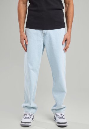 Jeans en denim bleu clair avec une coupe décontractée, présentant un design standard à cinq poches. Portés avec un t-shirt noir et des baskets blanches.