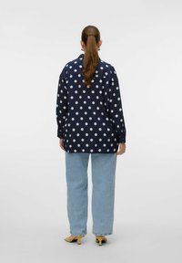 Chemise bleue marine à pois avec une coupe décontractée, des manches longues, des poignets boutonnés et un col. Associée à un jean bleu clair à jambes larges et des talons jaunes.