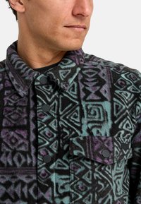 Chaqueta de felpa con patrones geométricos en negro, azul verdoso y púrpura. Cuenta con un cuello, botones a presión y un bolsillo en el pecho.