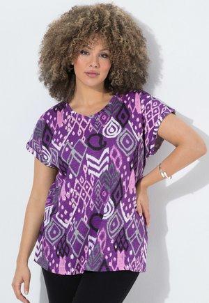 Frau mit lockigem Haar, die ein lila kurzärmliges Shirt mit abstrakten geometrischen Mustern, schwarze Hose und ein silbernes Armband trägt und mit der Hand an der Hüfte posiert.