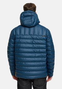 Blå pufferjakke med vandrette quiltede sektioner, en hætte og sort hardware. Har et logo på den øverste ryg. Båret over mørke jeans.