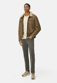 Braune Lederjacke mit Schaffellkragen, zwei Brusttaschen und Reißverschluss. Kombiniert mit einem cremfarbenen Hemd, grauen Jeans und beigen Sneakers.