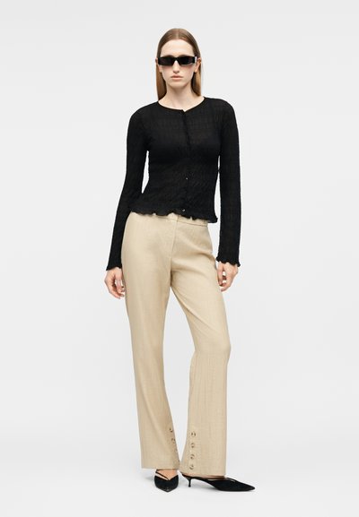 Γυναίκα που φοράει μαύρο υφασμάτινο μπλουζάκι με κουμπιά και μακριά μανίκια, μπεζ παντελόνι φαρδιάς γραμμής με κουμπιά στα μανσέτα, μαύρες μυτερές γόβες και μαύρα γυαλιά ηλίου.