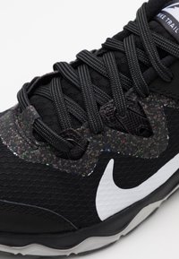 Chaussure de sport noire avec tissu texturé, accents multicolores tachetés sur la tige, et lacets noirs. Comprend un logo Nike swoosh blanc.