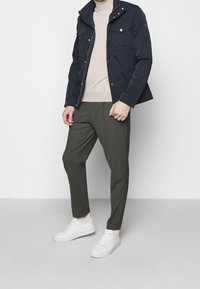 Námořnická bunda s několika kapsami, béžový svetr, šedé kalhoty a bílé tenisky. Jednoduchý, podaný střih s viditelnými detaily šití.