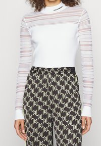 Haut blanc à manches longues avec des manches rayées texturées et transparentes, associé à un pantalon noir à motifs avec des dessins géométriques verts et blancs.