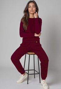 Bordeaux trui en joggingsbroek van zachte stof. De trui heeft een ronde halslijn, terwijl de joggingsbroek elastische boorden heeft. Witte sneakers maken de outfit compleet.