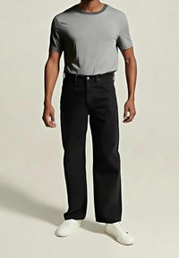 Homme portant un t-shirt gris à manches courtes, un pantalon noir droit et des chaussures blanches sans lacets, debout sur un fond uni.