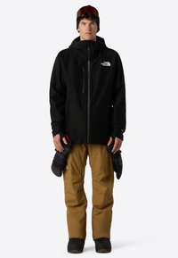 Sort vandtæt jakke med justerbar hætte, lynlåslommer og logo; brune isolerede bukser; handsker holdt i hænderne; vintertøj til snevejr.