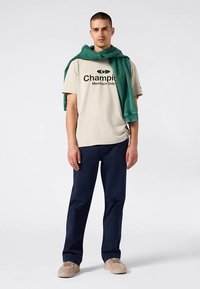 Camiseta de algodón beige con logo negro "Champion Members Only", pantalones azul marino y sudadera verde colgada sobre los hombros. Zapatos beige.