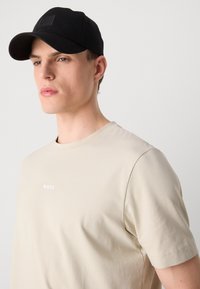 Νέος άνδρας φορώντας μια μπεζ μπλούζα BOSS και μια μαύρη καπέλο BOSS, κοιτώντας ελαφρώς προς τα αριστερά μπροστά σε απλό φωτεινό φόντο.