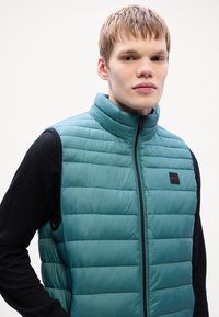 Gilet matelassé bleu sarcelle avec col haut, fermeture éclair sur le devant et chemise intérieure noire. Présente un petit logo de marque sur la poitrine gauche.