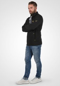 Sudadera negra con cuello alto, cordones blancos y acentos dorados. Llevada con jeans azules desgastados y zapatos blancos.