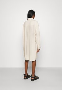 Beige übergroßes Hemdkleid mit Kragen, langen Ärmeln und Details an der Rückenpasse, kombiniert mit schwarzen Sandalen und glatter Hautstruktur.