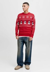 Suéter de punto rojo con diseños de muñecos de nieve, patrones de copos de nieve y un cuello acanalado. Llevado con jeans azules holgados y zapatos beige.
