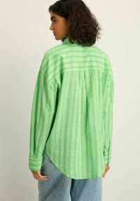 Camicia verde a righe con vestibilità comoda, dotata di maniche lunghe, piega sul retro e polsini con bottoni. Indossata con pantaloni in denim azzurro chiaro.