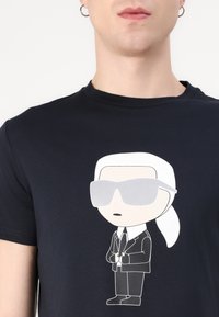 Juodas medvilnės marškinėliai su animaciniu personažu, apsirengusiu kostiumu ir akinių nuo saulės, šviesiai pilka ir balta grafika priekyje.