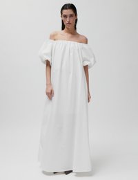 Γυναίκα με νωπά μαλλιά, φορώντας ένα λευκό μακρύ φόρεμα χωρίς ώμους με φουσκωτά μανίκια, στέκεται μπροστά σε απλό φόντο.