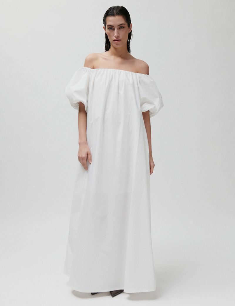 Γυναίκα με νωπά μαλλιά, φορώντας ένα λευκό μακρύ φόρεμα χωρίς ώμους με φουσκωτά μανίκια, στέκεται μπροστά σε απλό φόντο.