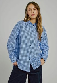 Blå rutiga skjortan med knäppning fram, har en spridd krage och svarta knappar, i kombination med mörka kordbyxor.