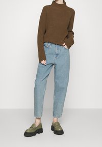 Person som bär en brun ribbad tröja, ljusblå hög midjejeans och olivgrön klumpig loafers, står framför en ljus bakgrund.