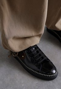 Sneaker din piele neagră, cu vârf rotund, șireturi frontale, cusături contrastante și un inel din piele maro. Purtați cu pantaloni călcati, lejere, de culoare bej.