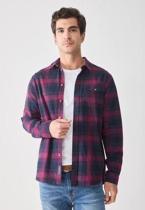 Camisa a cuadros en azul oscuro y burdeos con un bolsillo, cerrada con botones en la parte delantera y un cuello abierto. Lleva puesta una camiseta blanca con una tela texturada.