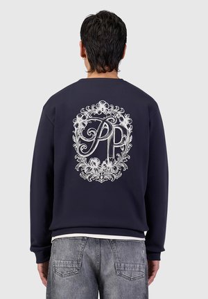 Persoon draagt een donker sweatshirt met een sierlijk wit bloemembleem en letters "PP" op de achterkant, gecombineerd met vervaagde blauwe spijkerbroek, met de rug naar voren.