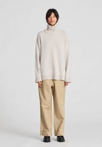 Vrouw die een beige coltrui, losse beige broek en zwarte schoenen draagt, staand tegen een effen lichtgrijze achtergrond.