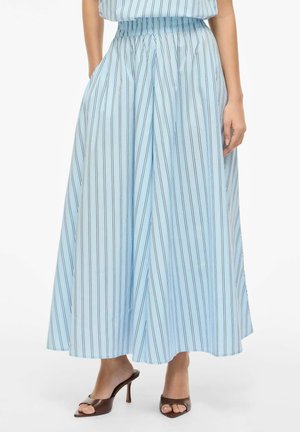 Naine, kes kannab helesinist kõrge vöökohaga, laiade säärtega triibulist seelikut taskutega ja pruune avatud varbaga kontsakingi.